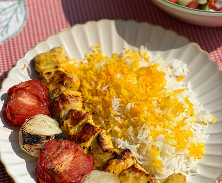 A delicious skewer of Joojeh KababPersian saffron chicken served with vibrant saffron rice, perfectly grilled tomatoes, and charred onions on a white plate.