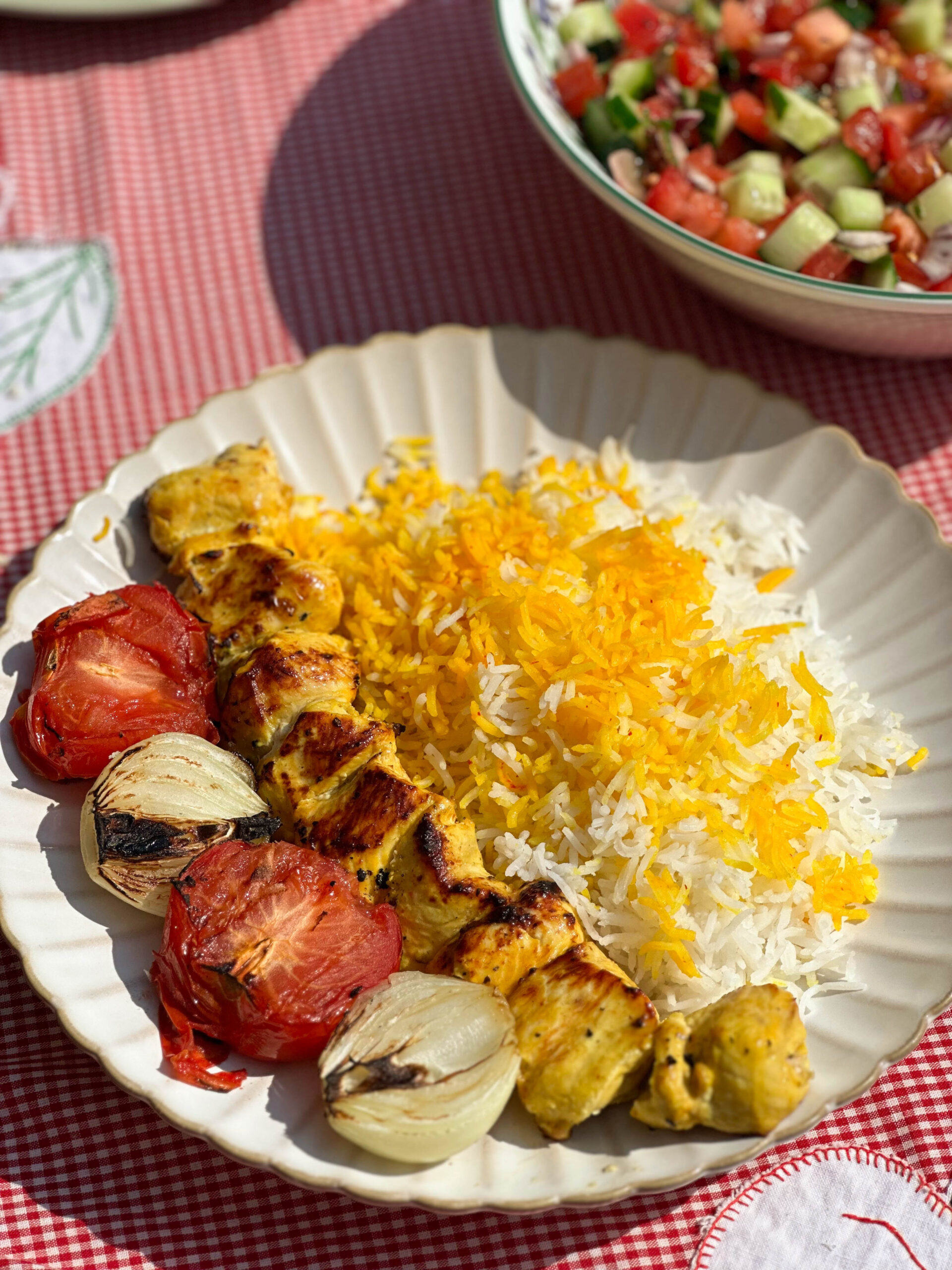 A delicious skewer of Joojeh KababPersian saffron chicken served with vibrant saffron rice, perfectly grilled tomatoes, and charred onions on a white plate.