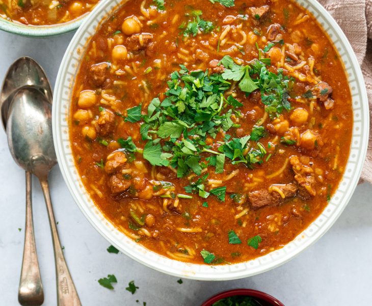 A bowl of steaming hot Authentic Moroccan Harira soup, filled with tender chunks of lamb, lentils, chickpeas, and aromatic spices, garnished with fresh parsley and a squeeze of lemon. A comforting and hearty dish that's perfect for a cold winter's day.