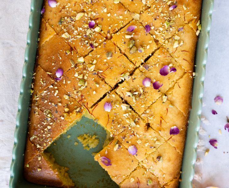 A close-up view of a delicious Pistachio Rose Baklava Cake. The cake is round and layered, with a golden brown crust. It is topped with a generous amount of crushed pistachios, giving it a vibrant green color. A drizzle of sweet rose syrup adds a touch of pink to the cake.