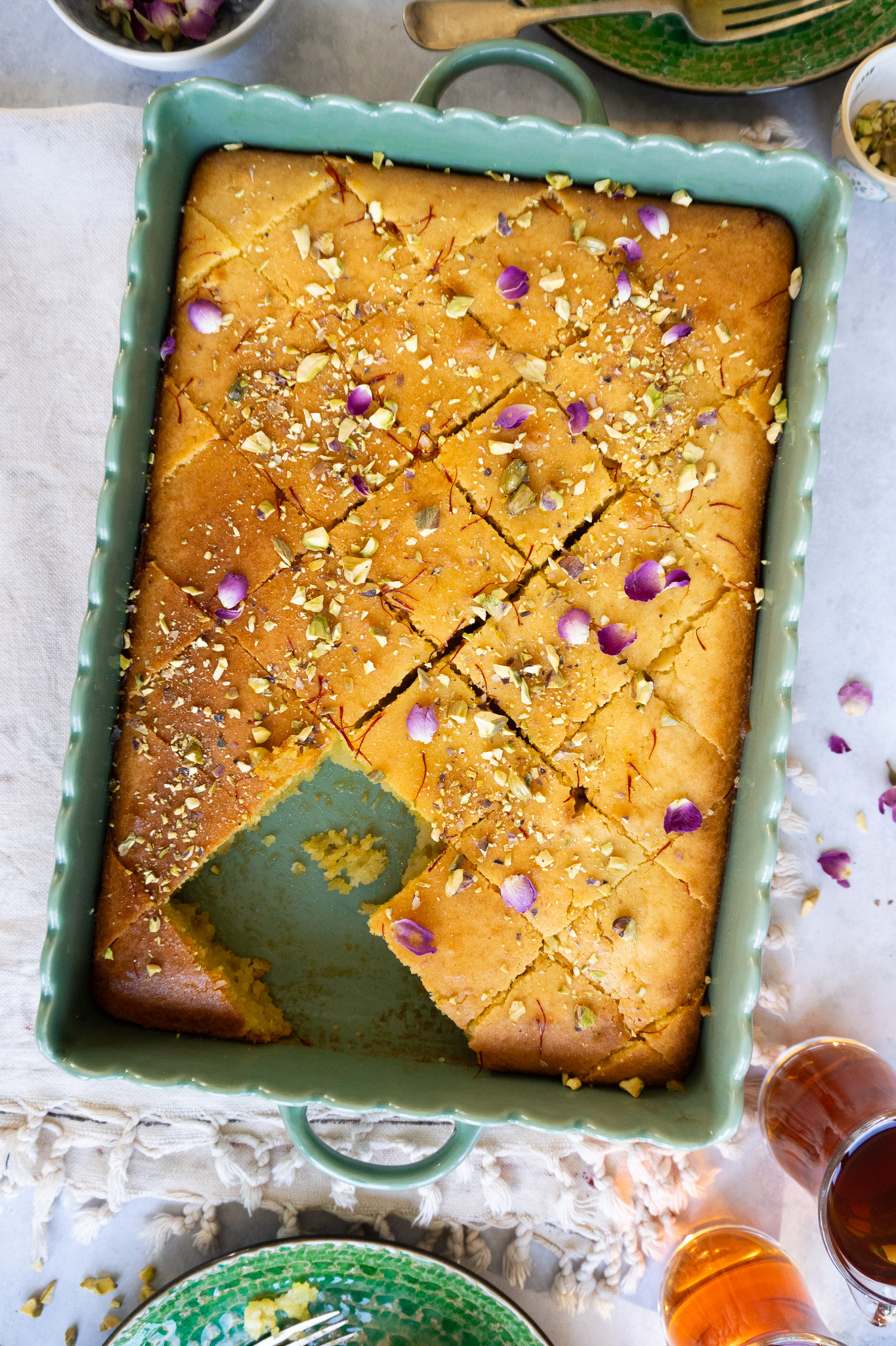 A close-up view of a delicious Pistachio Rose Baklava Cake. The cake is round and layered, with a golden brown crust. It is topped with a generous amount of crushed pistachios, giving it a vibrant green color. A drizzle of sweet rose syrup adds a touch of pink to the cake.