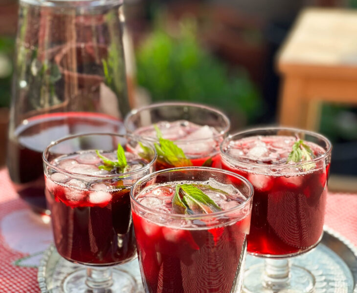 Glass of vibrant red Karkade - Hibiscus Tea on a tray, garnished with fresh mint leaves and ice cubes.